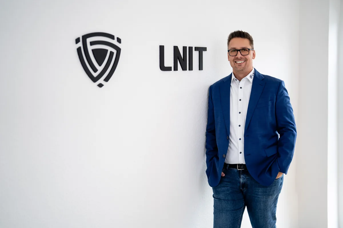 Lars Neumann im blauen Sakko lehnt lächelnd an der weißen Wand mit dem LNIT-Logo
